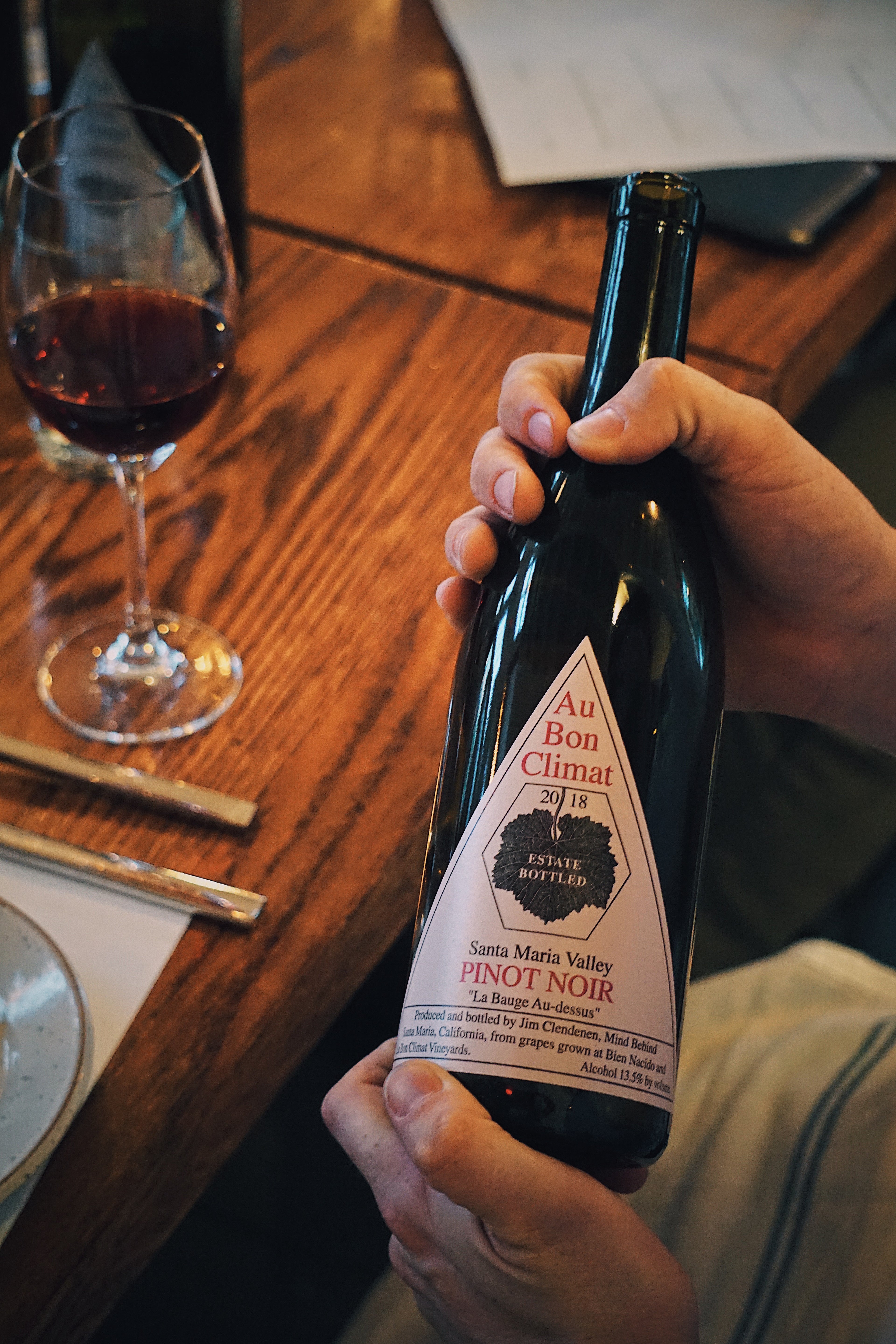 A close-up of Au Bon Climat Pinot Noir over a wooden dining table set with plates and a glass of red wine. 