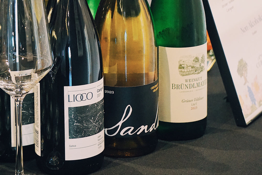 Collection of wine bottles (Lioco, Sandhi, Brundlmayer) on a table with a glass and a framed document in the background.