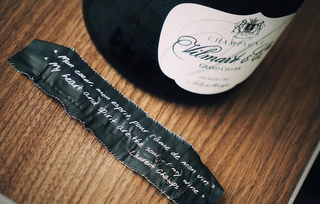 An artisanal bottle of Vilmart Champagne resting on a rustic wood surface next to its discarded foil, which has been smoothed out to show elegant French script on the interior.
